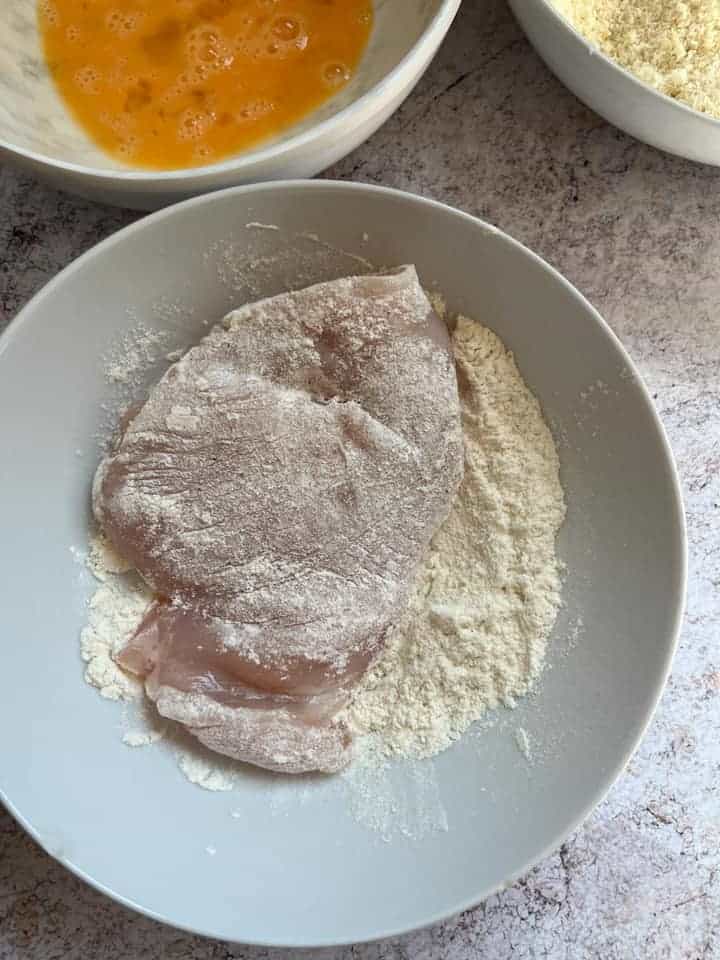 a chicken fillet in a white bowl of flour and a bowl of beaten egg.