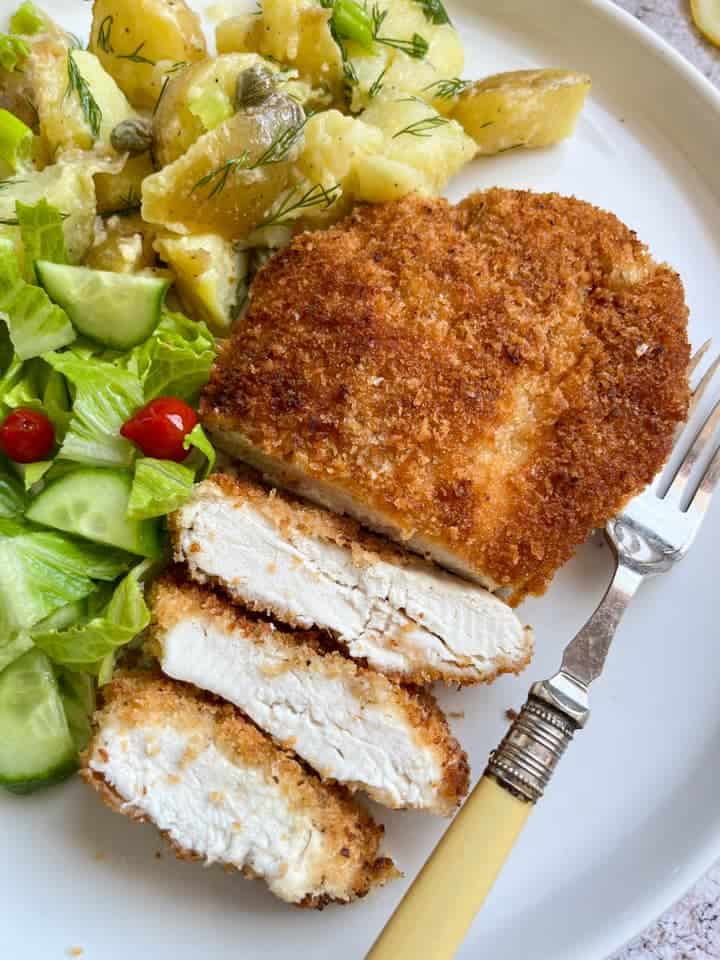 a golden breadcrumbed piece of chicken partially sliced on a white plate with potato salad, lettuce, cucumber and mini red peppers.