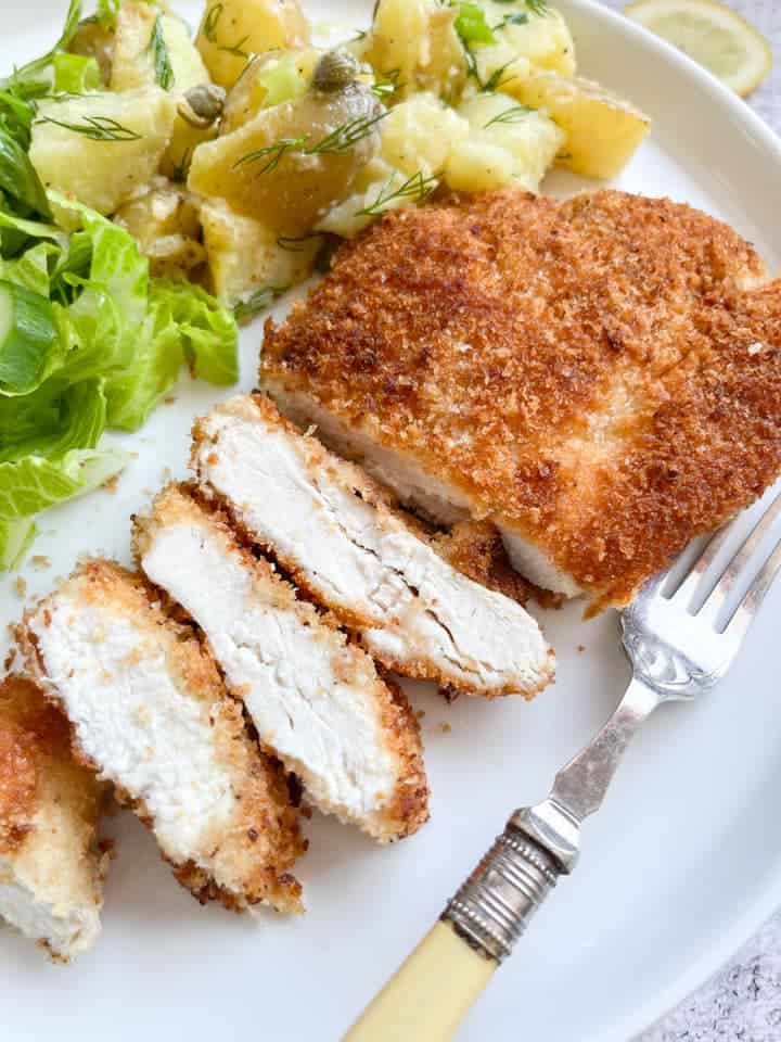 a partially sliced chicken Schnitzel in breadcrumbs on a white plate with potato salad with capers and lettuce. 