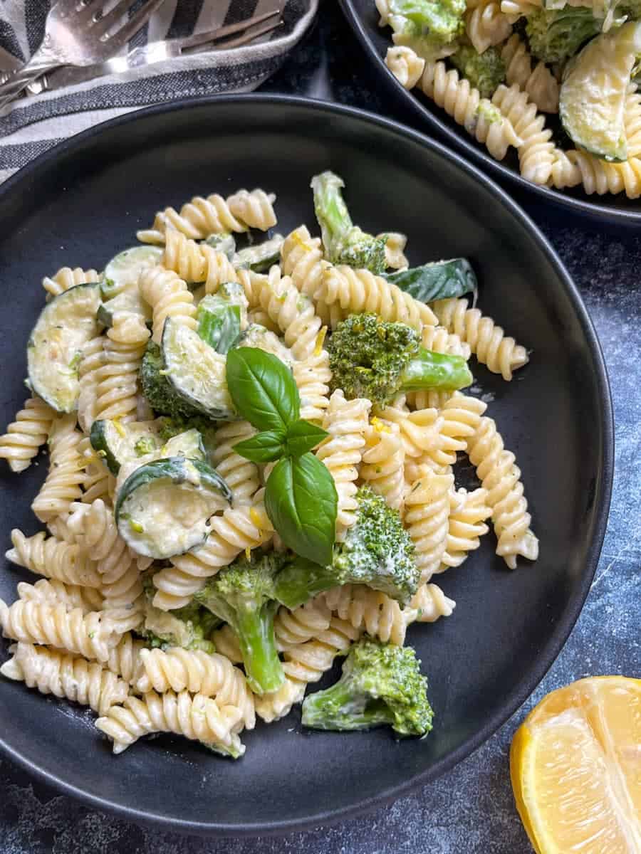 a black bowl of Fusilli pasta with sauteed zucchini and broccoli in a creamy garlic herb sauce topped with a fresh basil leaf.