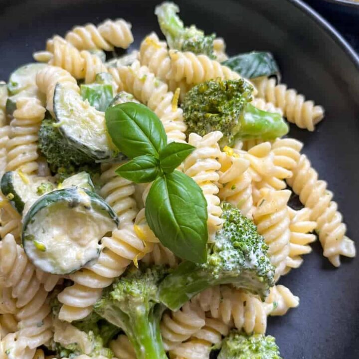 a black bowl of Fusilli pasta with sauteed courgettes and broccoli in a creamy garlic herb sauce topped with a fresh basil leaf.