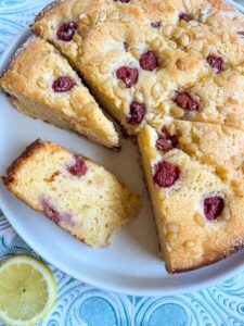 a partially sliced ricotta cake topped with raspberries and pine nuts on a white plate and a slice of lemon on a blue and white background.