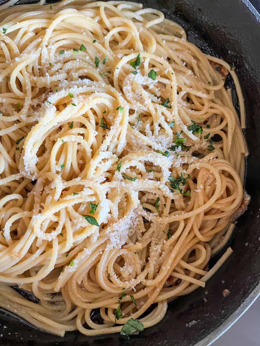 A black saucepan of spaghetti sprinkled with grated parmesan cheese and chopped parsley.