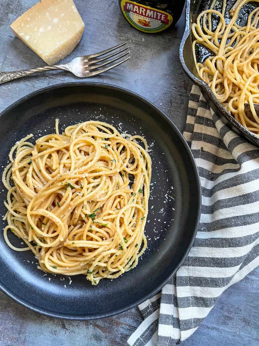 a black bowl of cooked spaghetti sprinkled with grated parmesan cheese and freshly chopped parsley, a silver fork, a black and white striped tea towel, a jar of marmite and a pan of spaghetti.