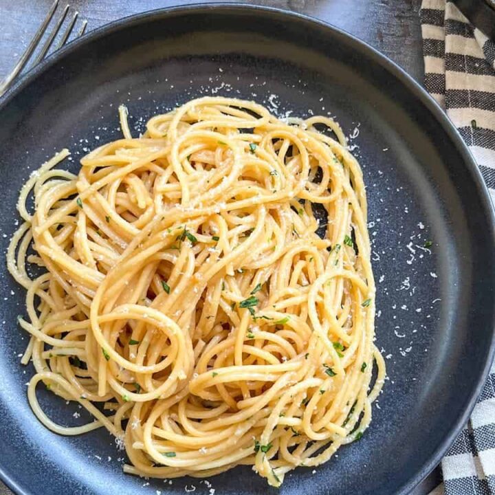 A black stoneware bowl of cooked spaghetti sprinkled with grated parmesan cheese and chopped parsley and a black and white striped tea towel.