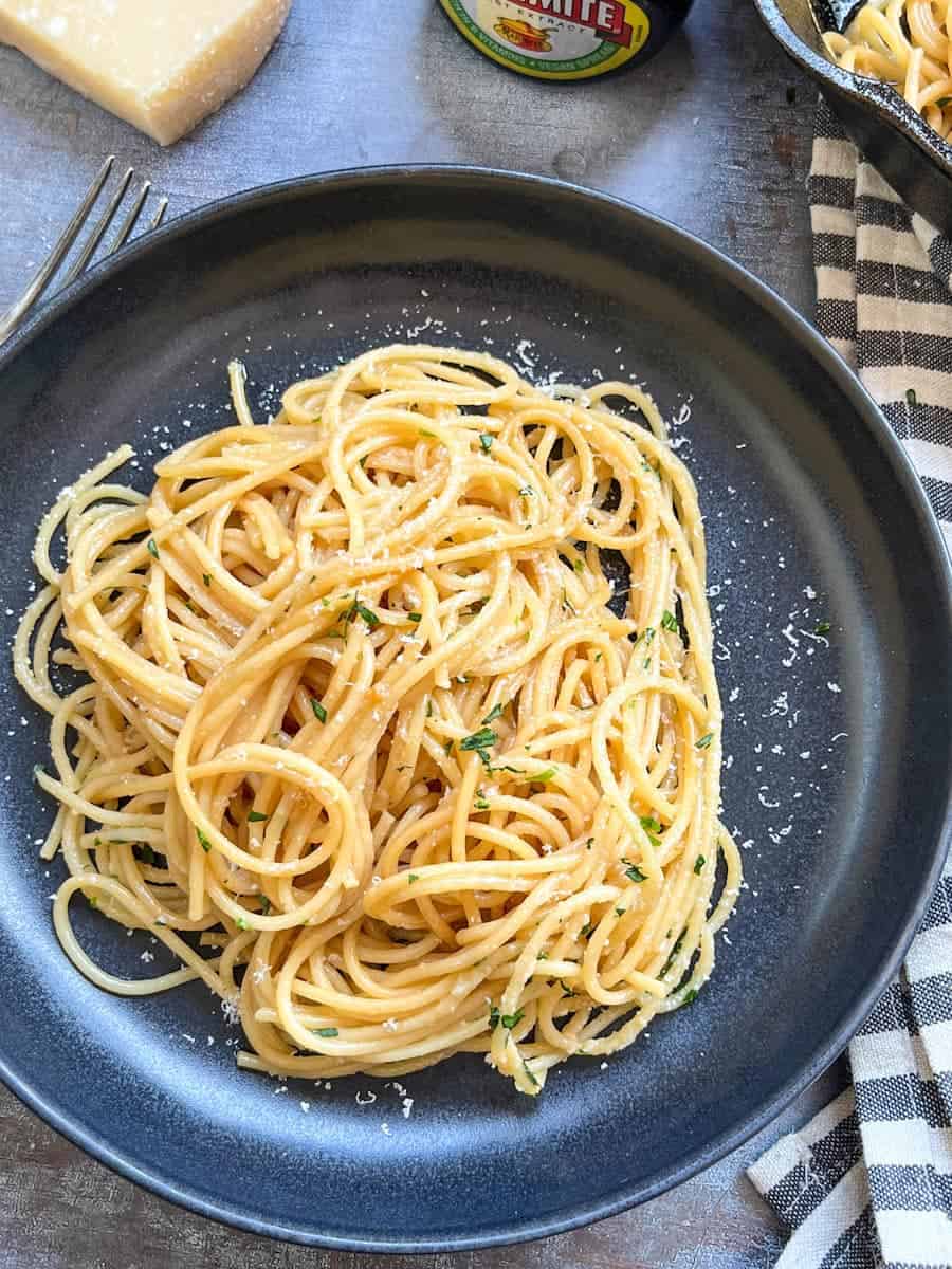 A black stoneware bowl of cooked spaghetti sprinkled with grated parmesan cheese and chopped parsley and a black and white striped tea towel.