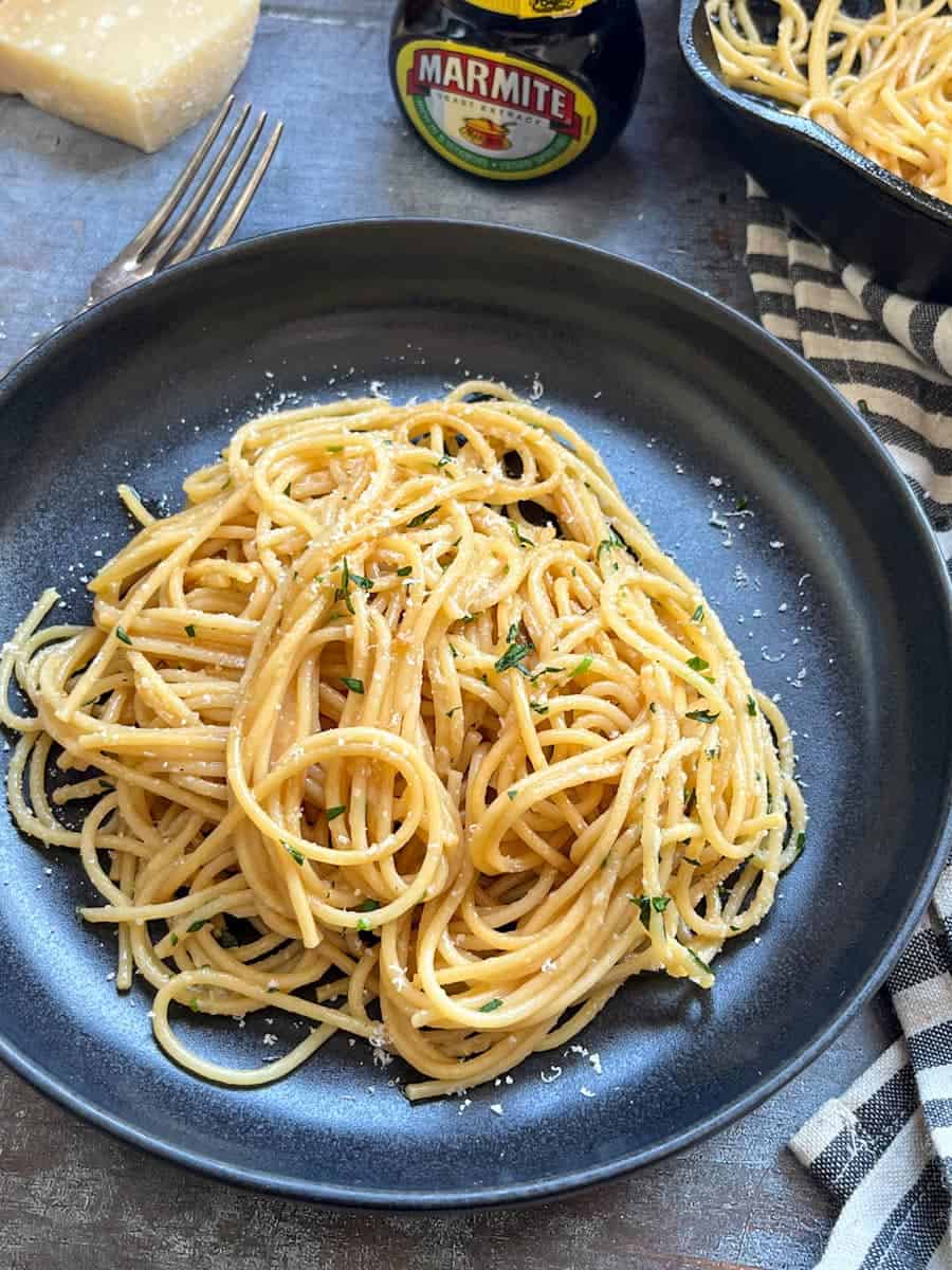 a black bowl of cooked spaghetti sprinkled with grated parmesan cheese and chopped parsley, a silver fork, a jar of marmite and a block of parmesan cheese.