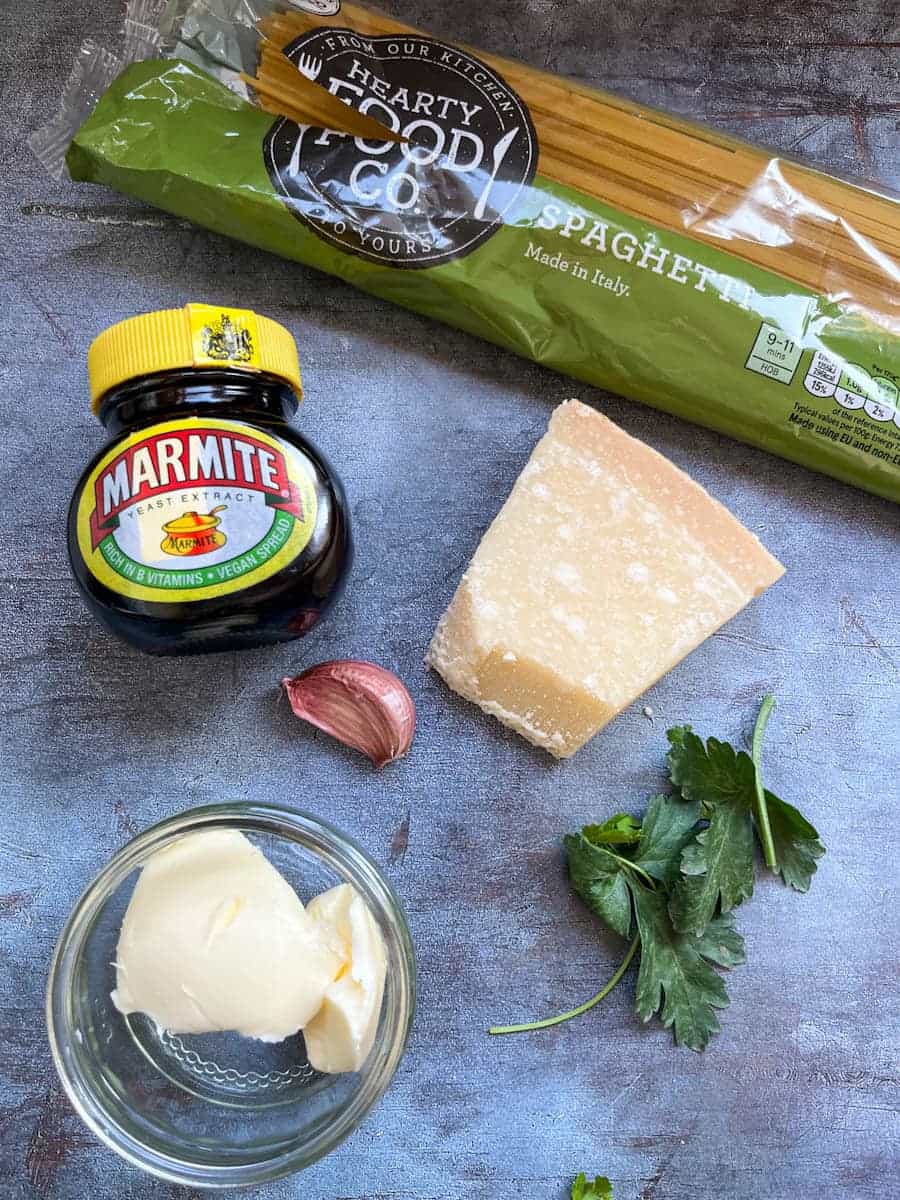 A packet of spaghetti, a block of parmesan cheese, a jar of marmite, a garlic clove, flat leaf parsley and a small jar of butter on a grey backdrop.