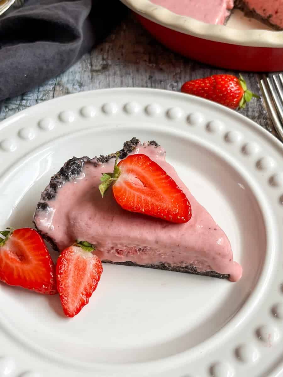 A slice of strawberry ice cream pie topped with a sliced fresh strawberry on a white plate.