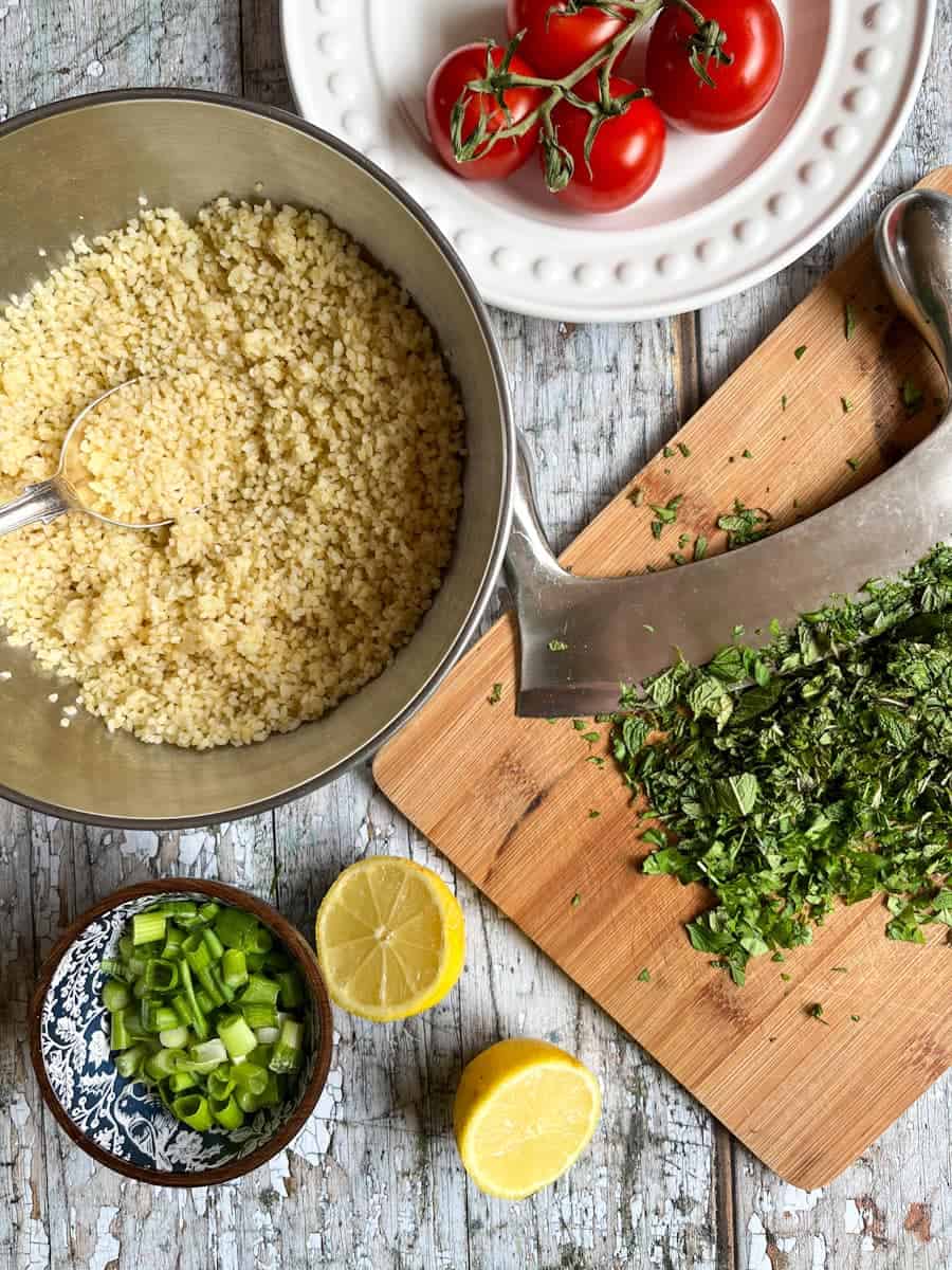 a silver bowl of bulgur wheat with a silver spoon, chopped fresh herbs on a chopping board, a white plate of tomatoes, a blue wooden bowl of chopped spring onions and a lemon cut in half.