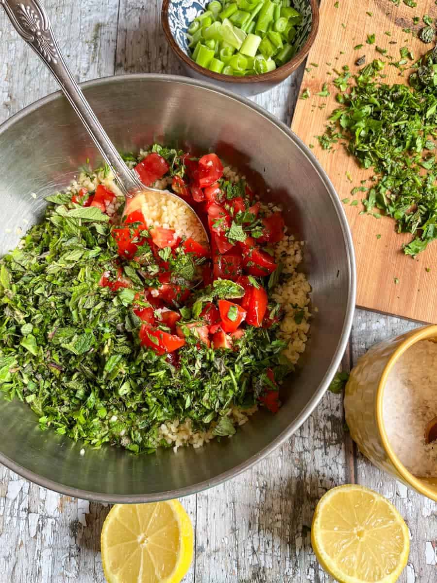 a silver bowl of  bulgur wheat, chopped tomatoes and fresh herbs with a silver spoon, a blue wooden bowl of spring onions, a yellow pot of salt, a lemon cut in half and a wooden chopping board with chopped herbs.