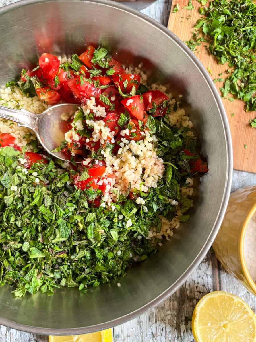 a silver bowl of bulgur wheat with diced tomatoes and fresh chopped herbs with a silver spoon.