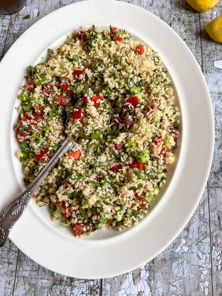 A Bulgur wheat salad with chopped tomatoes, fresh herbs and spring onions on a white plate with a silver spoon.