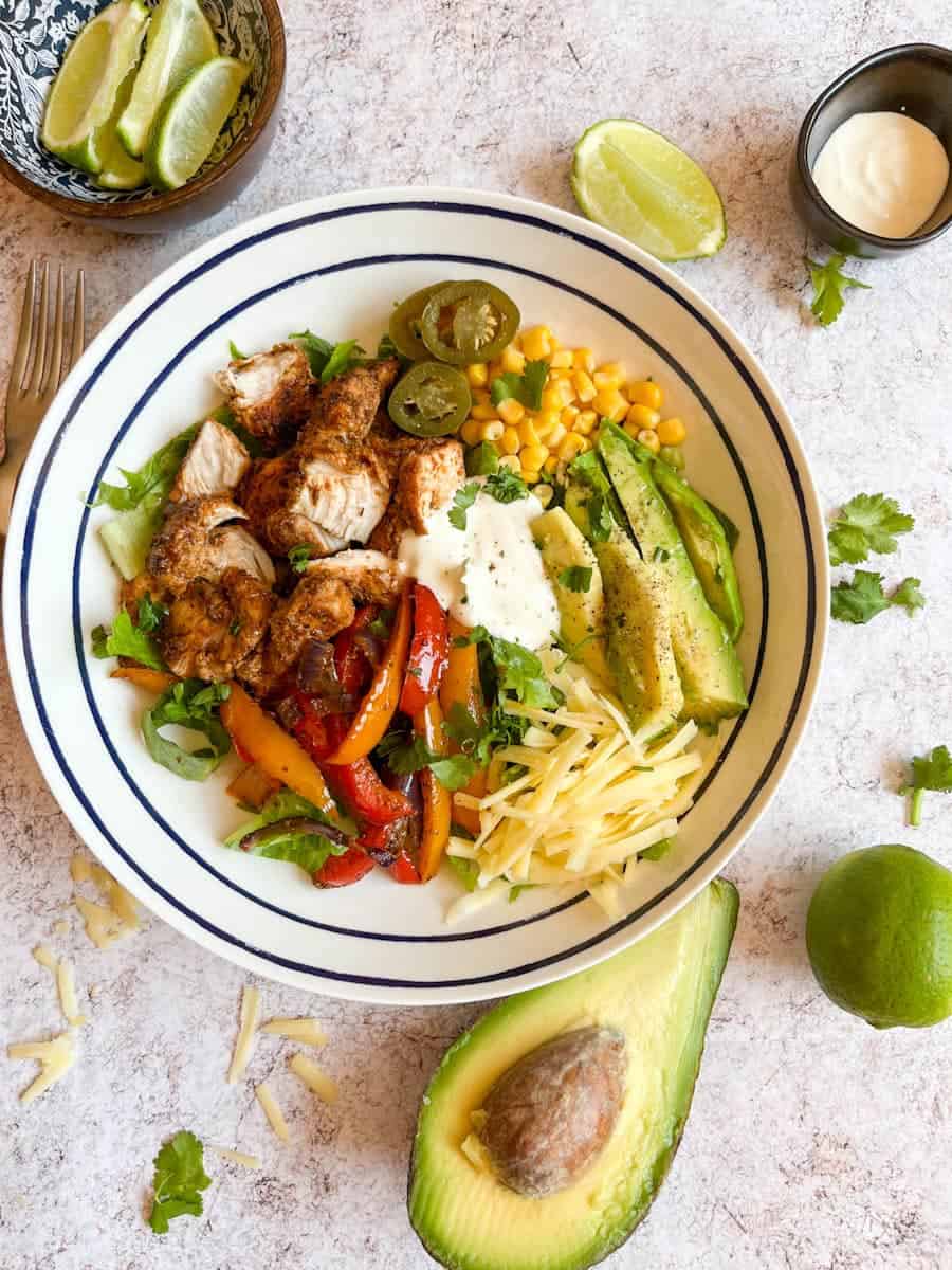 a blue and white bowl of cooked Fajita chicken pieces, shredded lettuce, grated cheese, avocado, sweetcorn, jalapenos and red and orange peppers. A bowl of lime wedges, a black pot of sour cream, coriander leaves and half an avocado are placed beside the bowl.