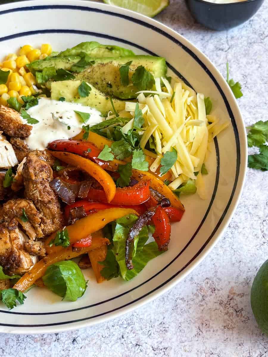 a blue and white bowl of chicken chunks in Fajita spice with onions, peppers, shredded lettuce, avocado, sweetcorn, grated cheese and sour cream.