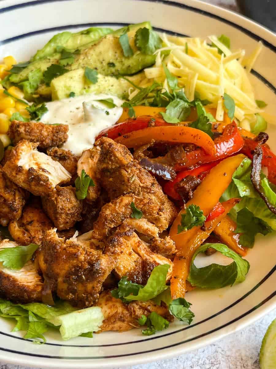 A blue and white bowl of Fajita spiced chicken chunks with onions, peppers, grated cheese, shredded lettuce and sour cream.