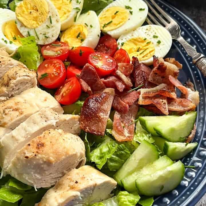 A cobb salad on a blue plate with cooked chicken, bacon pieces, cucumber, tomatoes, hard boiled eggs and lettuce scattered with fresh chives.