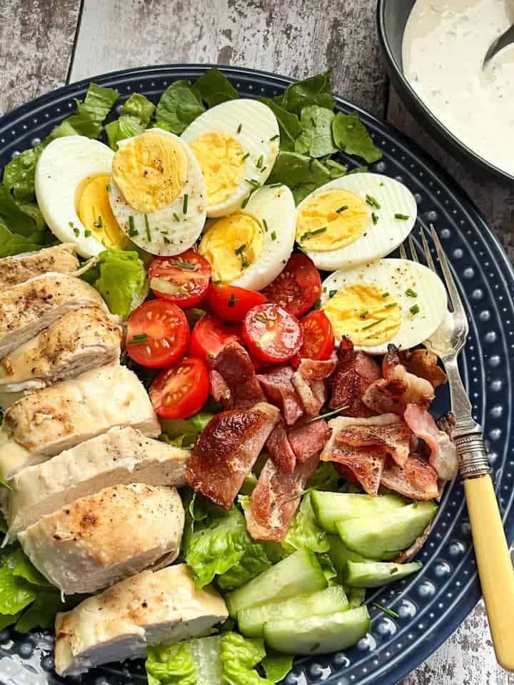 A salad with sliced grilled chicken, hard boiled eggs, cooked bacon pieces, sliced cucumber, tomatoes and crispy green lettuce on a blue plate with a bowl of creamy herb dressing.