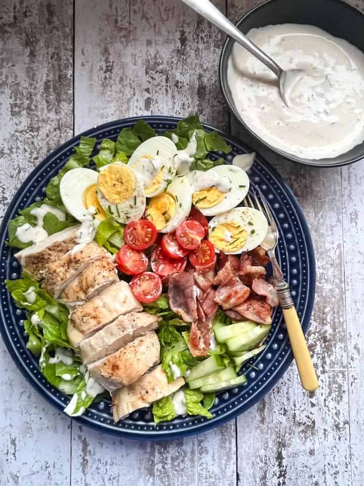 A salad with grilled sliced chicken, crispy bacon, hard boiled eggs, sliced tomatoes, cucumber and lettuce drizzled with a creamy dressing on a blue plate.  A bowl of the salad dressing with a serving spoon sits above the plate. 