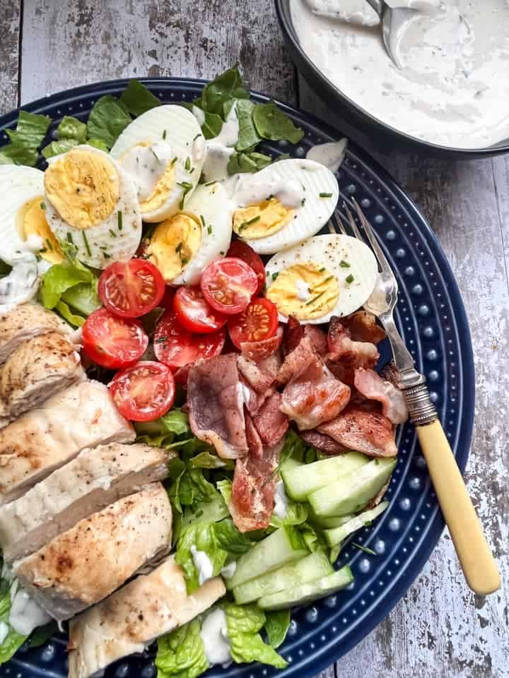 A salad with sliced grilled chicken, lettuce, crispy bacon, hard boiled eggs, tomatoes and sliced cucumber on a blue plate with a bowl of creamy herb dressing on the side. 