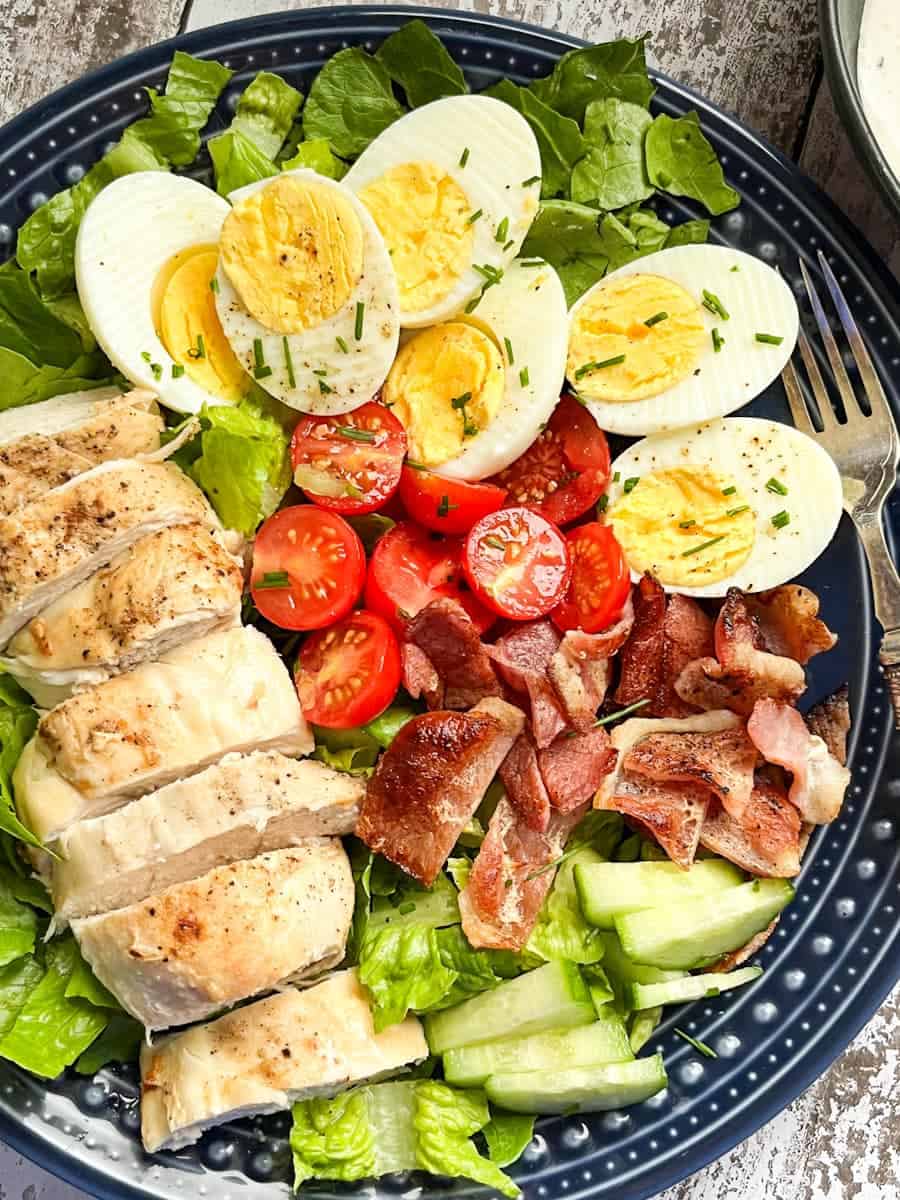 A salad with grilled chicken, cooked bacon lettuce, tomatoes, cucumber and hard boiled eggs sprinkled with chopped chives on a blue plate.