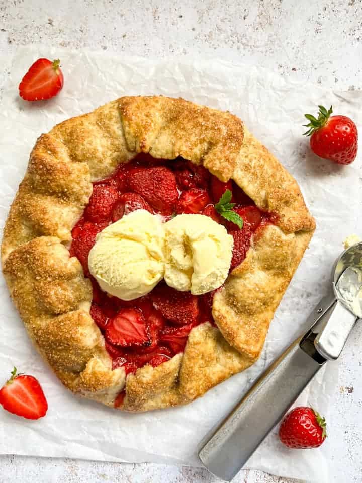 A freshly baked Galette filled with strawberries topped with two scoops of vanilla ice cream and a fresh sprig of mint on a piece of baking parchment. A silver ice cream scoop and fresh strawberries are beside the galette.