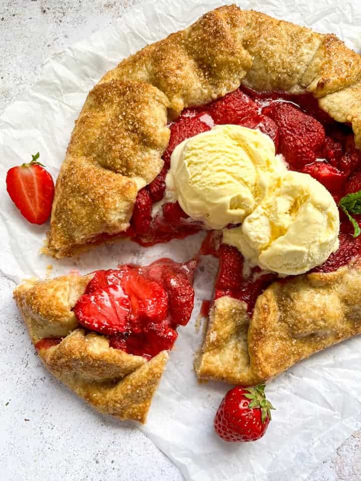 A strawberry Galette topped with two scoops of vanilla ice cream on a piece of baking paper. A slice of the Galette has been cut and the strawberry filling is running out onto the paper.