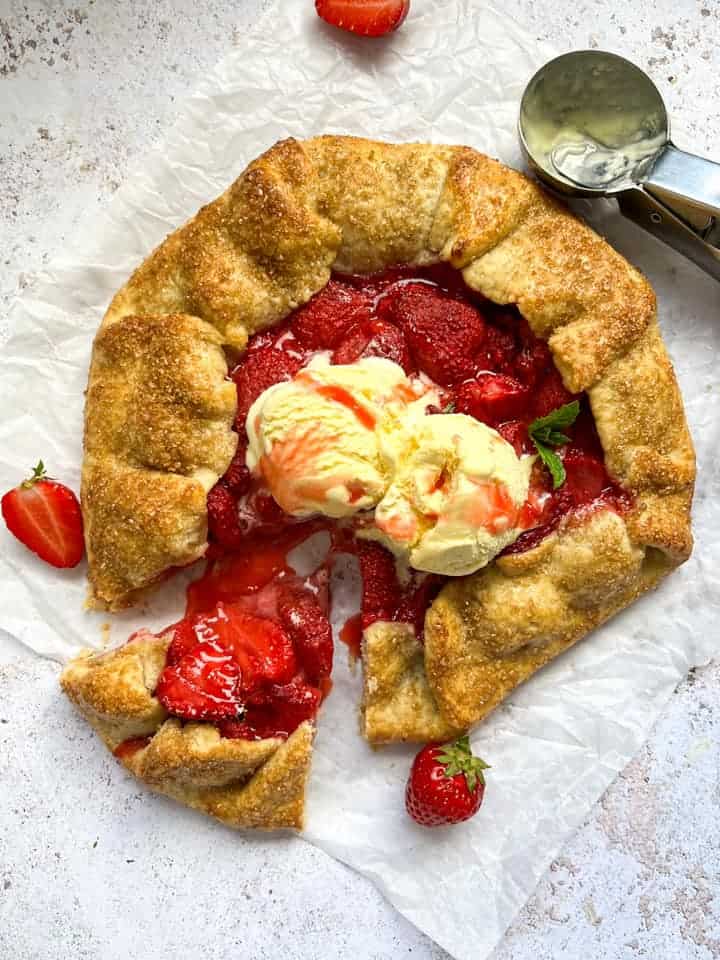 A galette filled with fresh strawberries topped with vanilla ice cream on a piece of baking parchment. A slice has been cut from the Galette and the strawberry filling is running onto the parchment. A silver ice cream scoop sits above the galette.