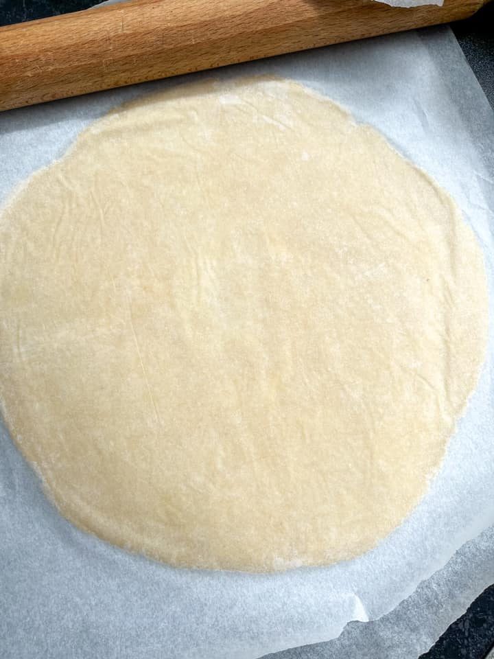 A circle of pie dough rolled out between two large sheets of baking parchment and a wooden rolling pin.