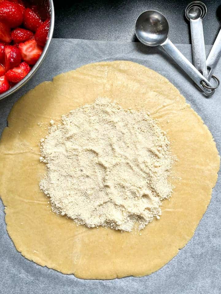A large circle of rolled out pie dough sprinkled with ground almonds, a set of silver measuring spoons and a silver bowl of strawberries.