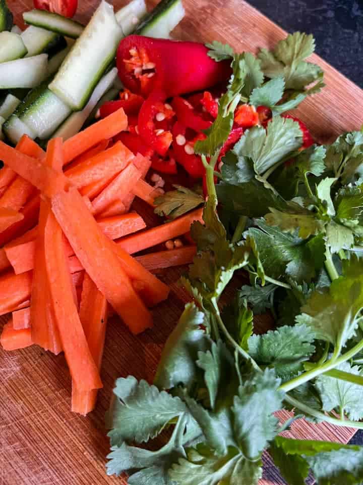 finely sliced carrot, cucumber, sliced red chilli and a bunch of fresh coriander on a wooden chopping board.