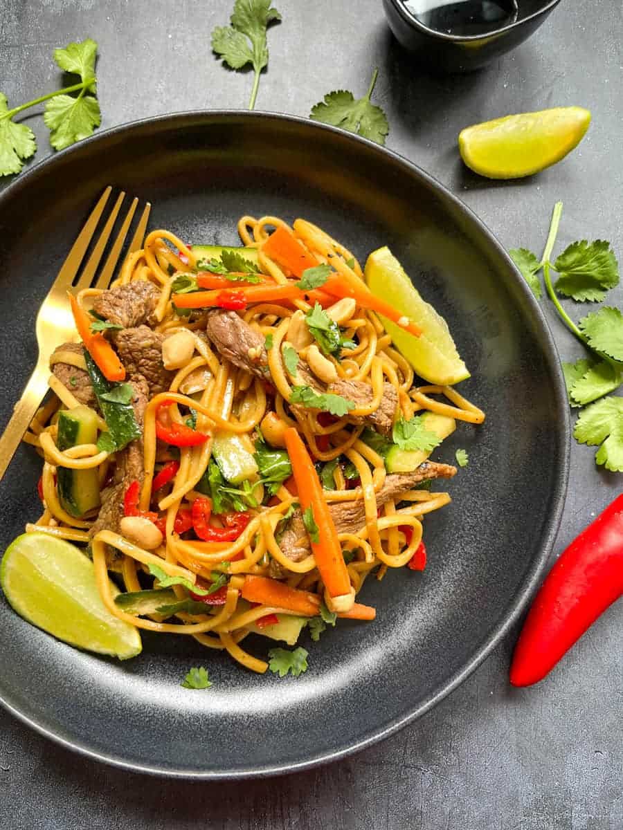 A beef noodle salad with sliced beef, sliced cucumber and carrot, egg noodles, fresh coriander and peanuts with lime wedges on a black plate with a gold fork.