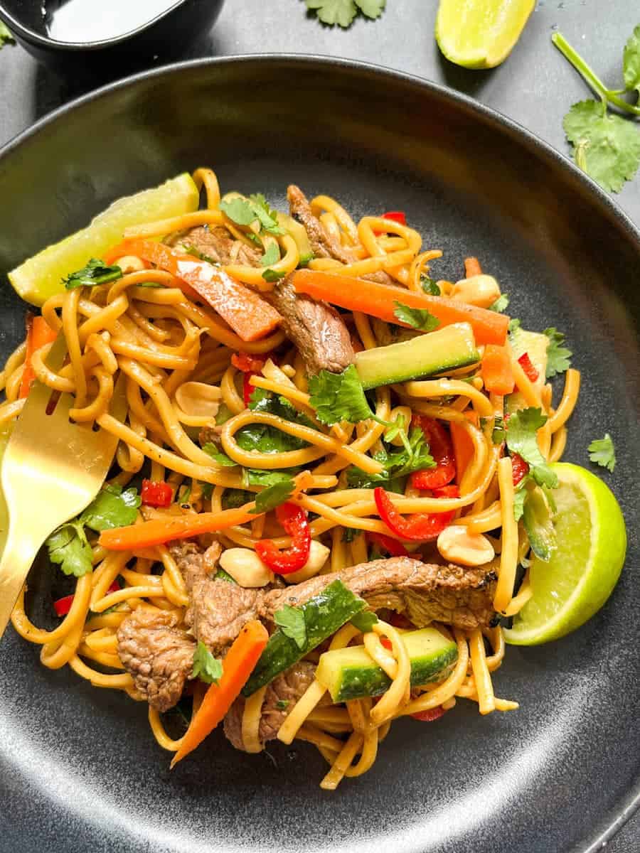 A beef noodle salad with sliced beef, noodles, sliced cucumber and carrot, sliced red chilli and fresh coriander with a lime wedge on a black plate with a gold fork.