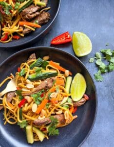 Two black bowls of noodles with sliced steak, sliced cucumber and carrot and red chilli with fresh coriander and peanuts.