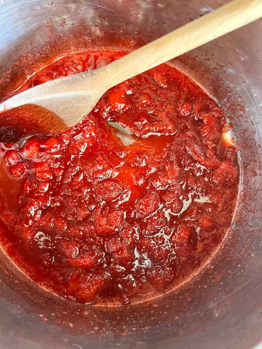 Strawberry compote cooking in a silver pan with a wooden spoon.