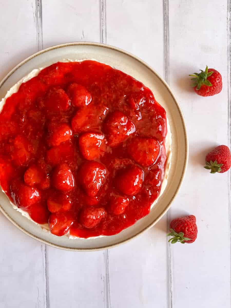 A cheesecake topped with strawberry compote on a white and beige plate on a wooden backdrop.