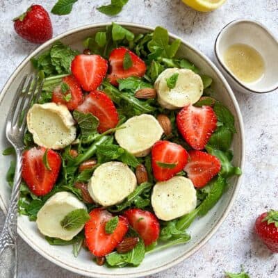 A rocket salad in a beige bowl with grilled goat's cheese slices, fresh strawberries, mint leaves and almonds with a silver fork resting against the bowl. A pot of lemon salad dressing, a halved lemon, strawberries and mint leaves sit beside the bowl.