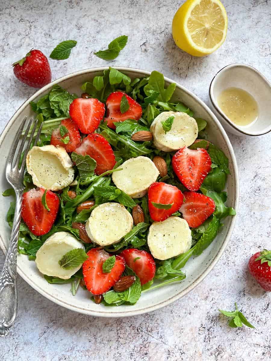 A rocket salad in a beige bowl with grilled goat's cheese slices, fresh strawberries, mint leaves and almonds with a silver fork resting against the bowl. A pot of lemon salad dressing, a halved lemon, strawberries and mint leaves sit beside the bowl.