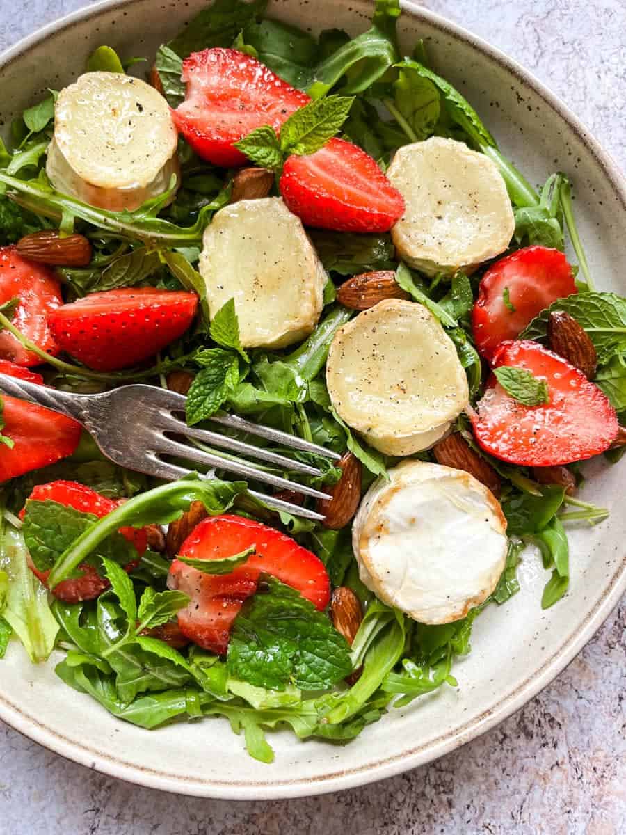 A rocket salad in a beige bowl with grilled goat's cheese slices, halved strawberries, fresh mint leaves and whole almonds with a silver fork.