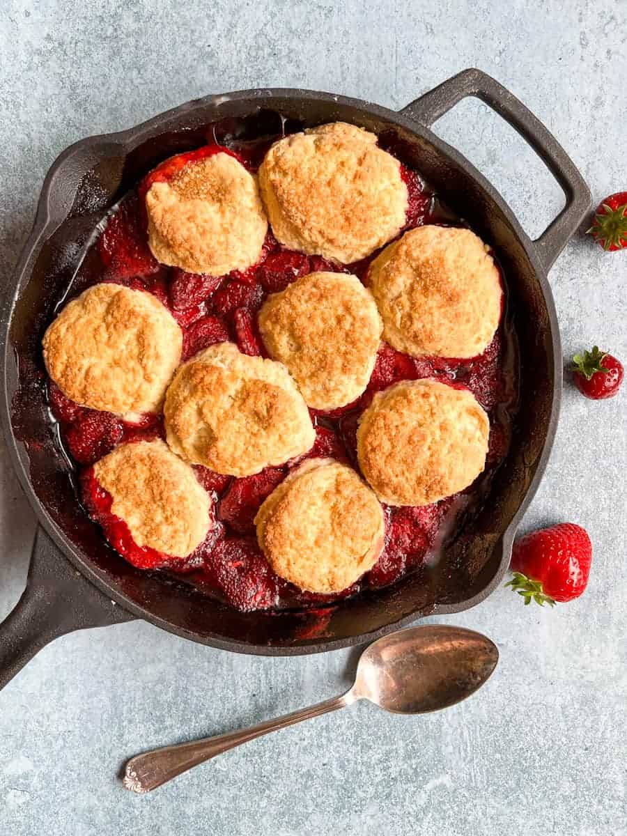 A strawberry cobbler in a black skillet with a silver spoon and fresh strawberries.