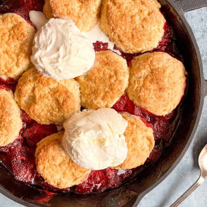 A strawberry cobbler topped with two scoops of vanilla ice cream in a black cast iron skillet.
