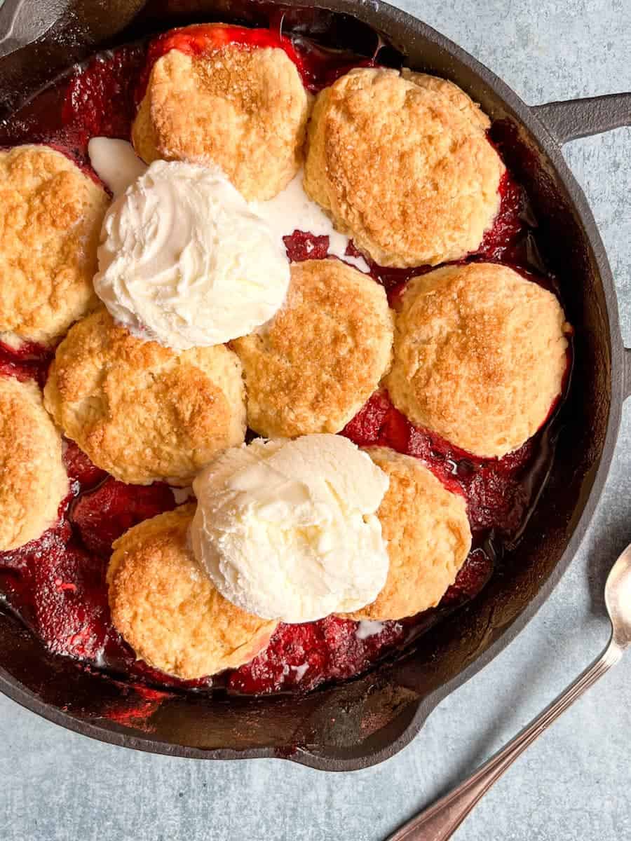 A strawberry cobbler topped with two scoops of vanilla ice cream in a black cast iron skillet.