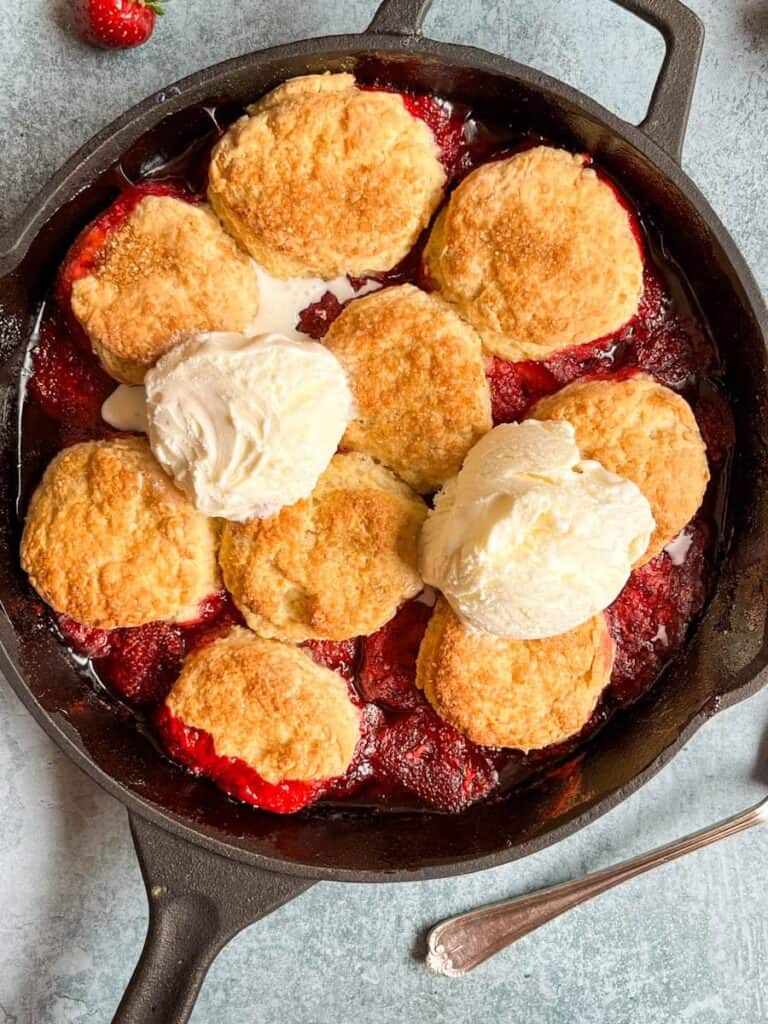 A strawberry cobbler topped with two scoops of vanilla ice cream in a black cast iron skillet.