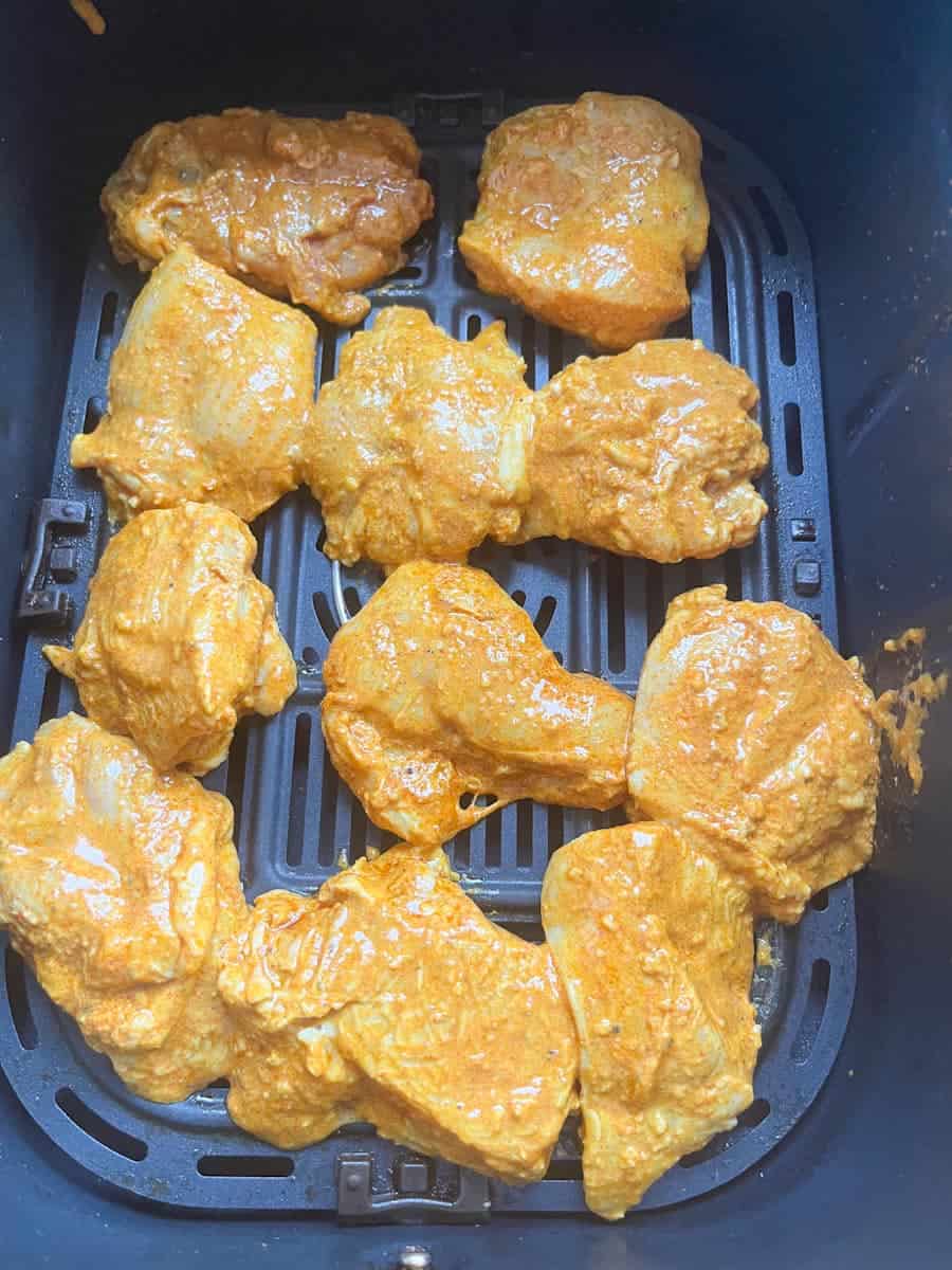 Raw chicken thigh pieces in a spicy yoghurt sauce in an air fryer basket.