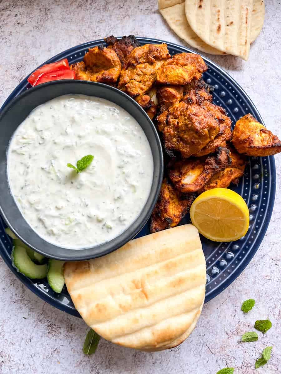 A blue plate of spicy chicken thigh pieces, a halved lemon, pitta bread, sliced cucumber and a black bowl of cucumber and mint dip.