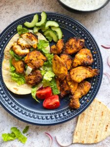 A blue plate of spiced chicken thigh pieces with pitta bread, shredded lettuce, tomatoes and cucumbers.