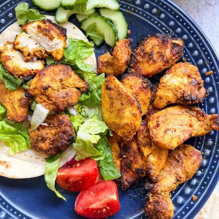 A blue plate of spiced chicken pieces with pitta bread, shredded lettuce, tomatoes and sliced cucumber.