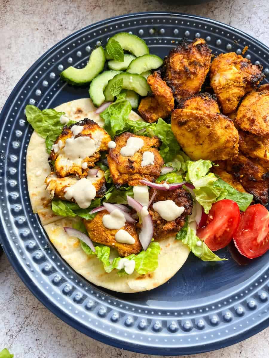 A blue plate of spiced chicken pieces with pitta bread, shredded lettuce, red onion, tomatoes and garlic sauce.