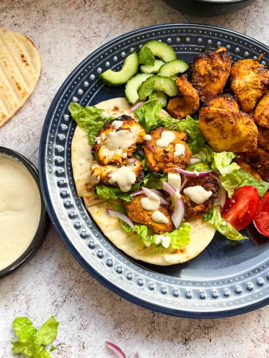 A blue plate of spiced chicken with pitta bread, shredded lettuce, sliced cucumbers, tomatoes, sliced red onion and garlic sauce.