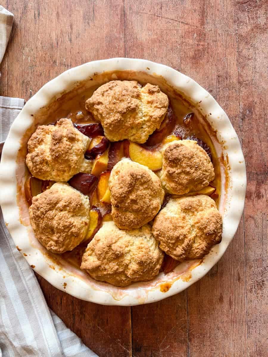 A peach cobbler in a cream round baking dish on a wooden background. 